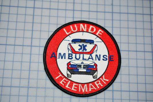 a red and white patch with the text "LUNDE AMBULANCE TELEMARK" and an illustration of an ambulance.