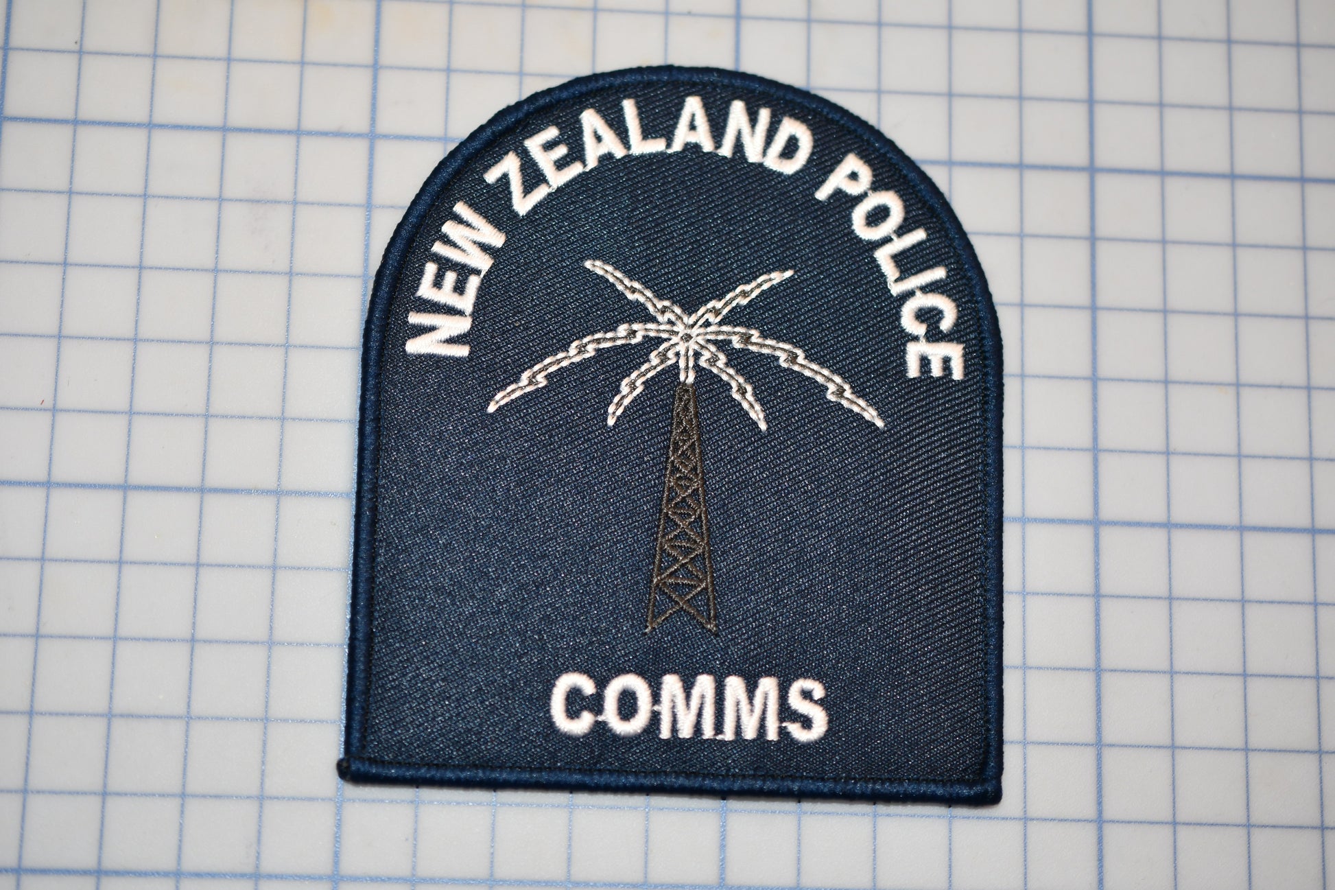 a New Zealand Police patch with a palm tree and a radio tower, along with the text "NEW ZEALAND POLICE" and "COMM".
