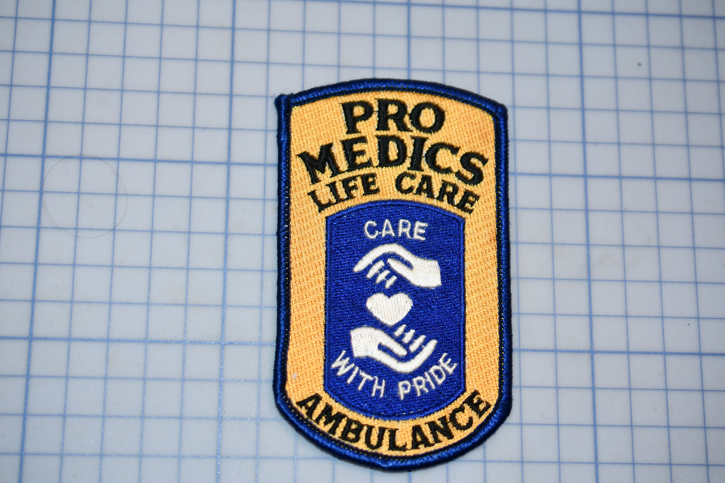 a patch with the text "PRO MEDICS LIFE CARE" and "CARE WITH PRIDE AMBULANCE" printed on it.