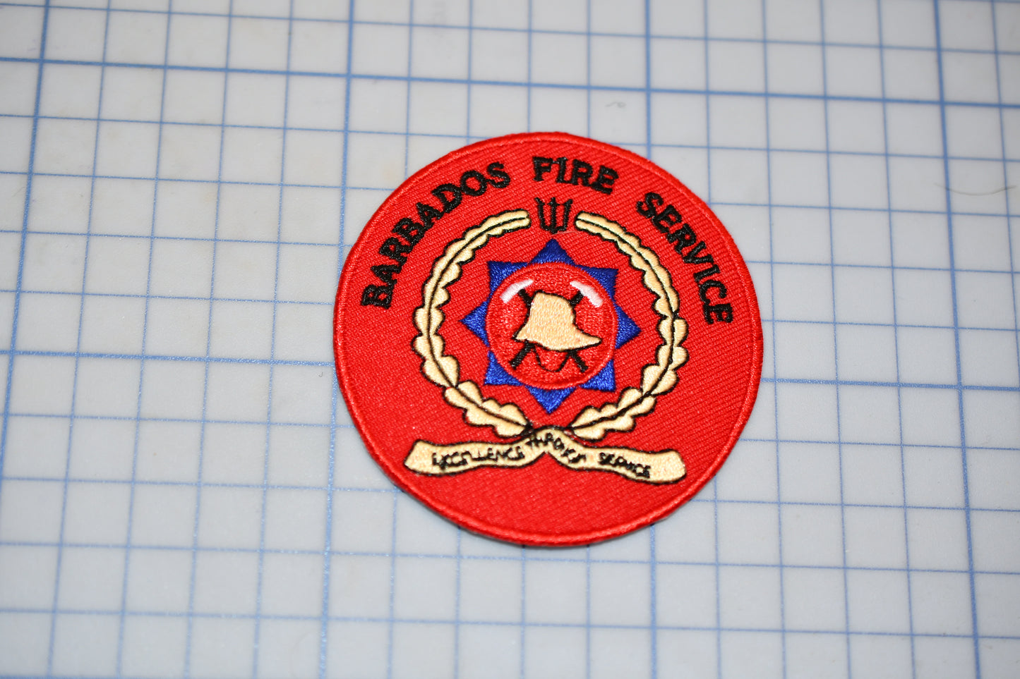 A red circular patch with the text "BARRADOS FIRE SERVICE" and a red emblem featuring a bell and a star, placed on a grid background.