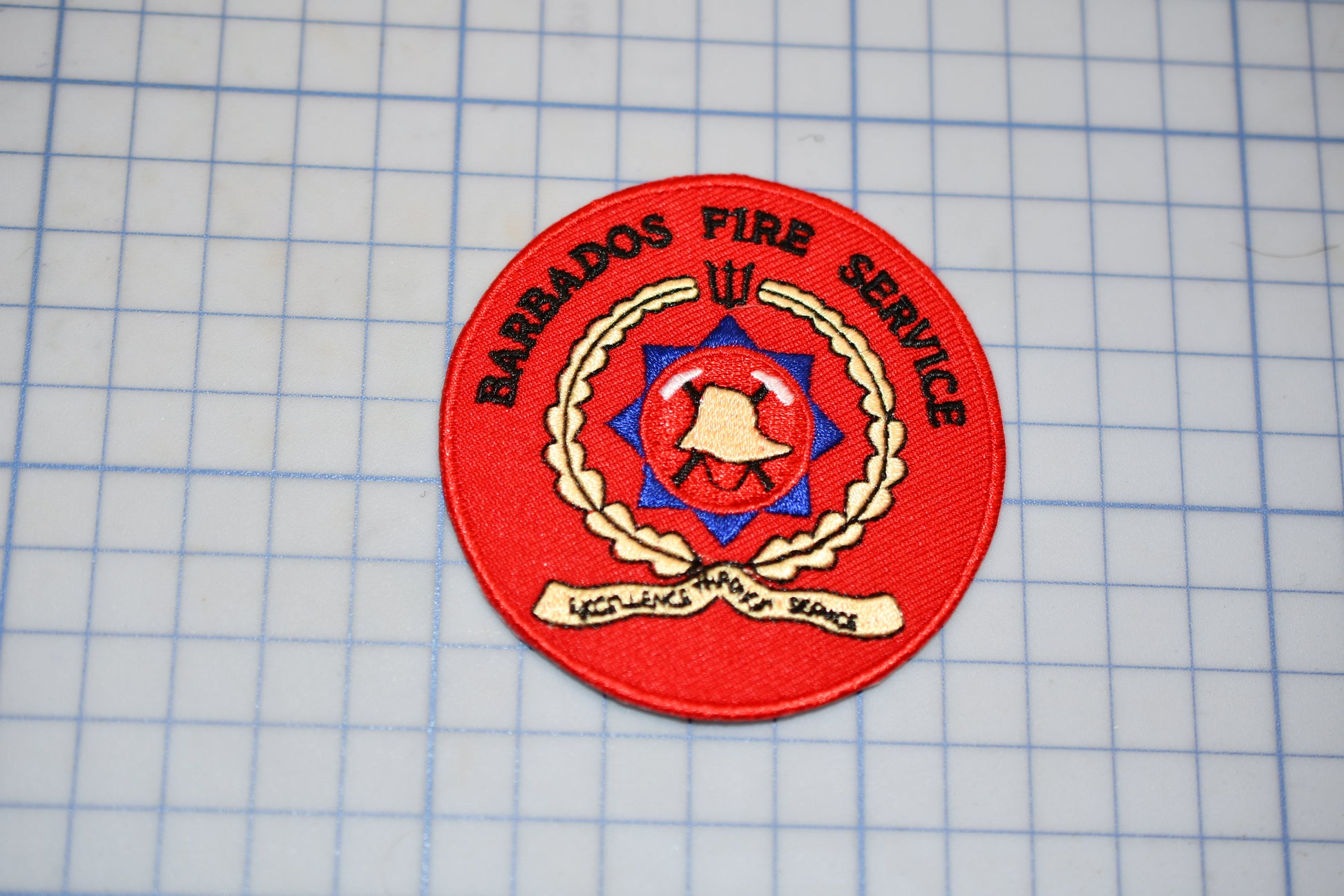 A red circular patch with the text "BARRADOS FIRE SERVICE" and a red emblem featuring a bell and a star, placed on a grid background.