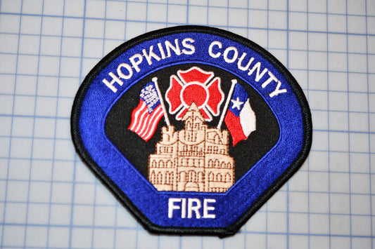 A blue and black patch with the text "HOPKINS COUNTY FIRE" and an illustration of a building with a fire hydrant and fire truck.