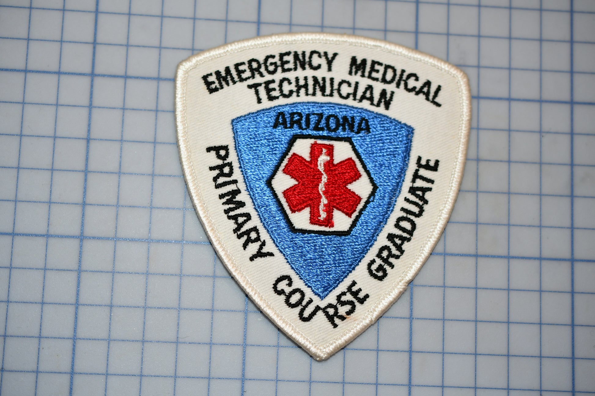 a patch with the text "EMERGENCY MEDICAL TECHNICIAN ARIZONA PRIMARY GRADUATE" and a red cross symbol, indicating that it is a patch for an emergency medical technician.