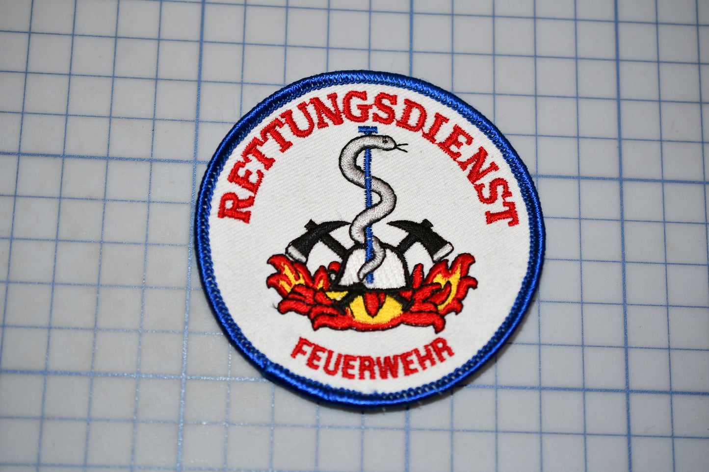 a circular patch with a red and blue design, including a snake, crossed swords, and a flame, surrounded by a blue border.