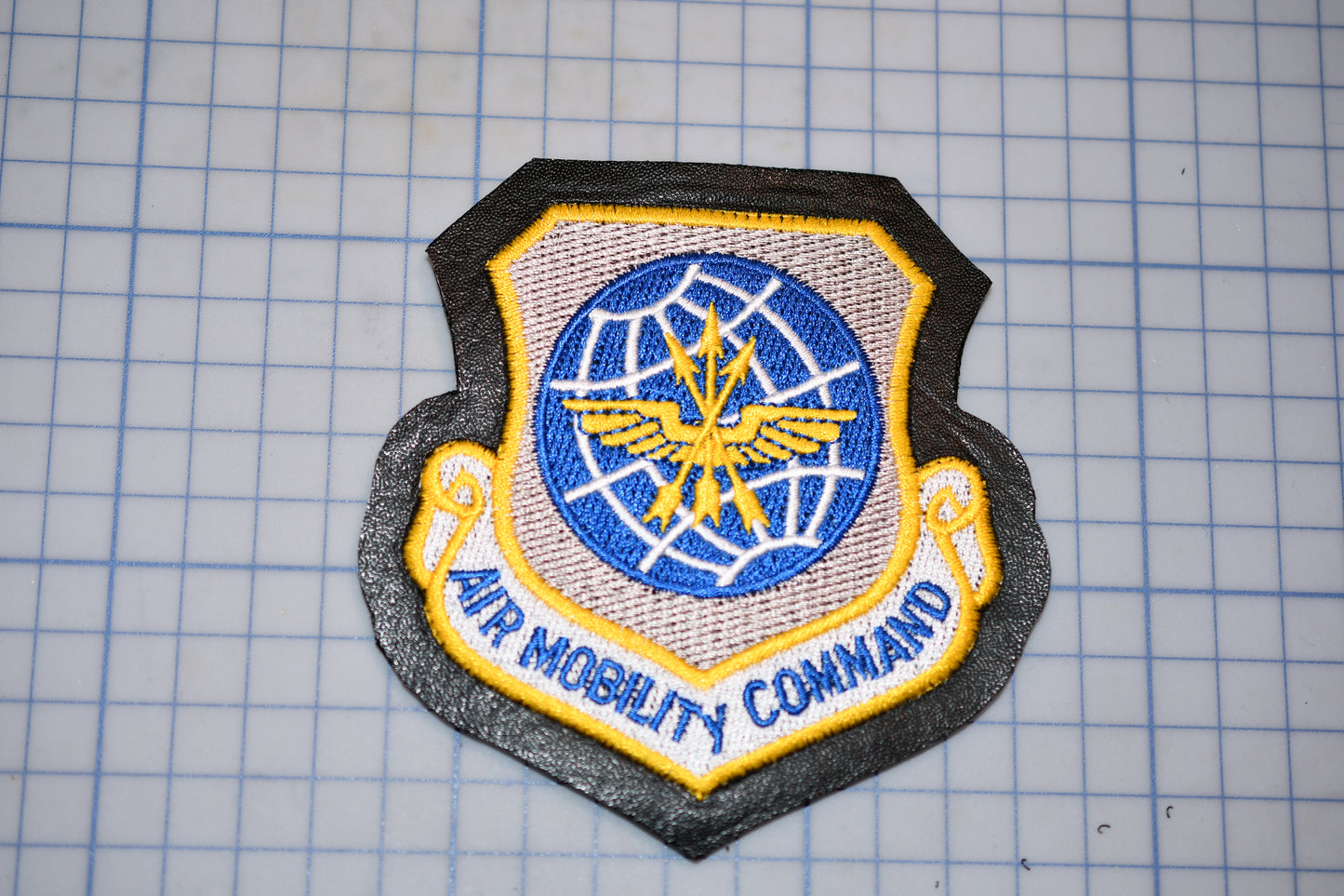 a military patch with a shield-shaped design, featuring a blue and yellow emblem with wings and a globe. The text "AIR MOBILITY COMMAND" is written below the emblem.