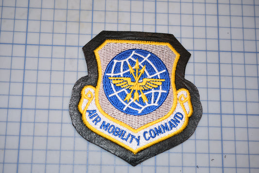 a military patch with a shield-shaped design, featuring a blue and yellow emblem with wings and a globe. The text "AIR MOBILITY COMMAND" is written below the emblem.