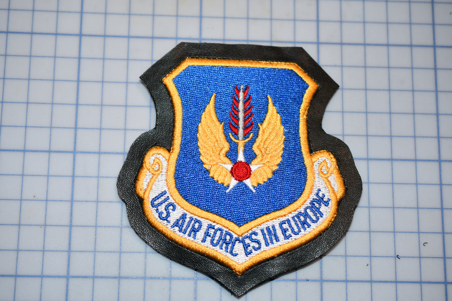 a U.S. Air Force patch with a blue background, yellow wings, and a red circle. The patch is placed on a grid background.