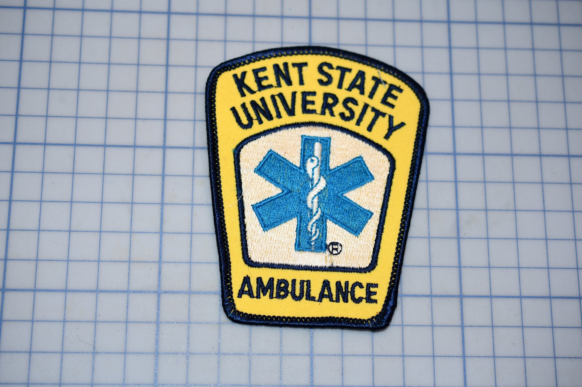 a yellow and blue patch with the text "KENT STATE UNIVERSITY AMBULANCE" and a medical symbol, indicating that it is a patch representing the Kent State University Ambulance Service.
