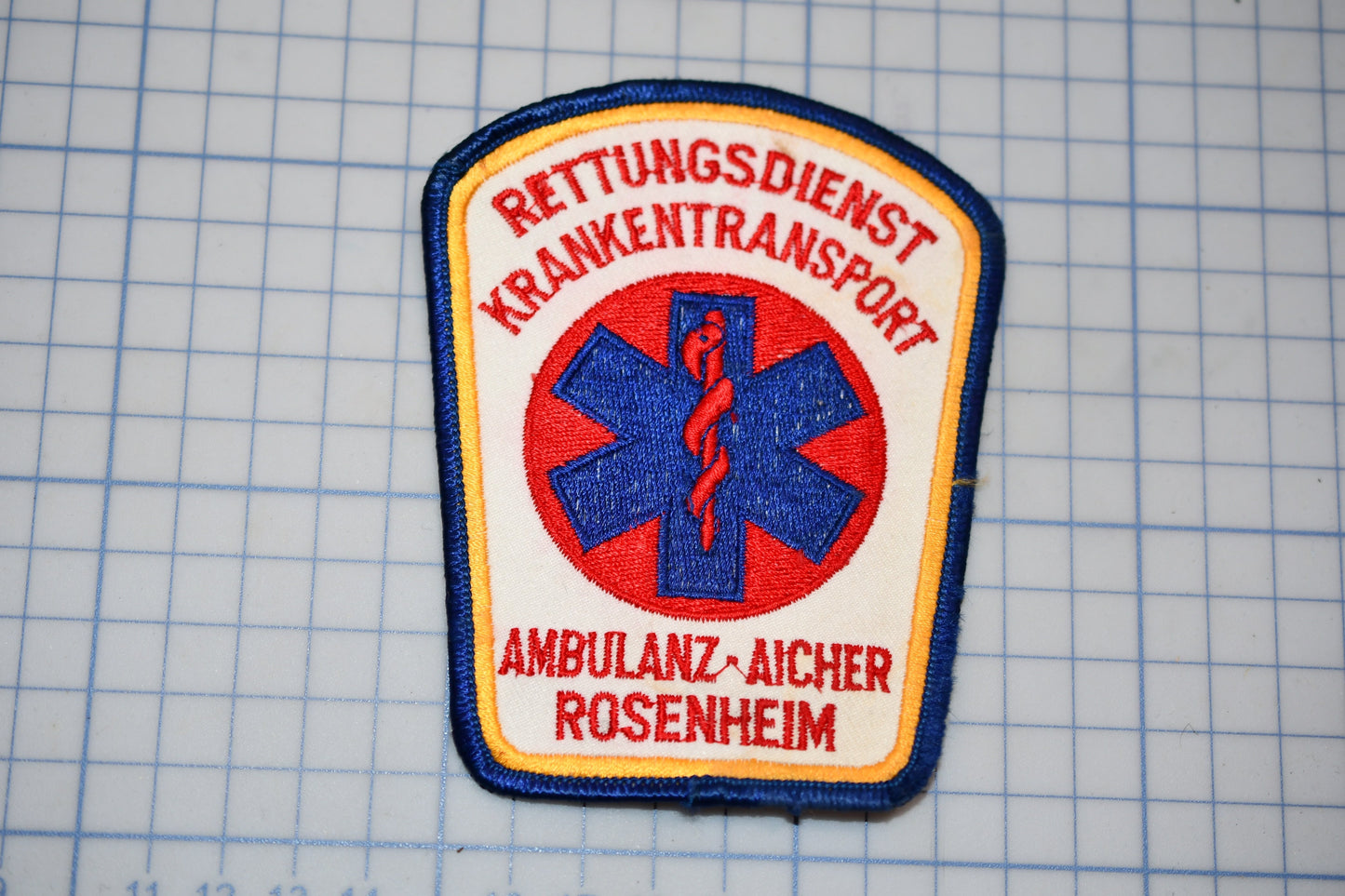 a patch with the text "Rettungsdienst Krankenttransport" and "Ambulanz-Aicher Rosenheim" printed on it, along with a medical symbol.