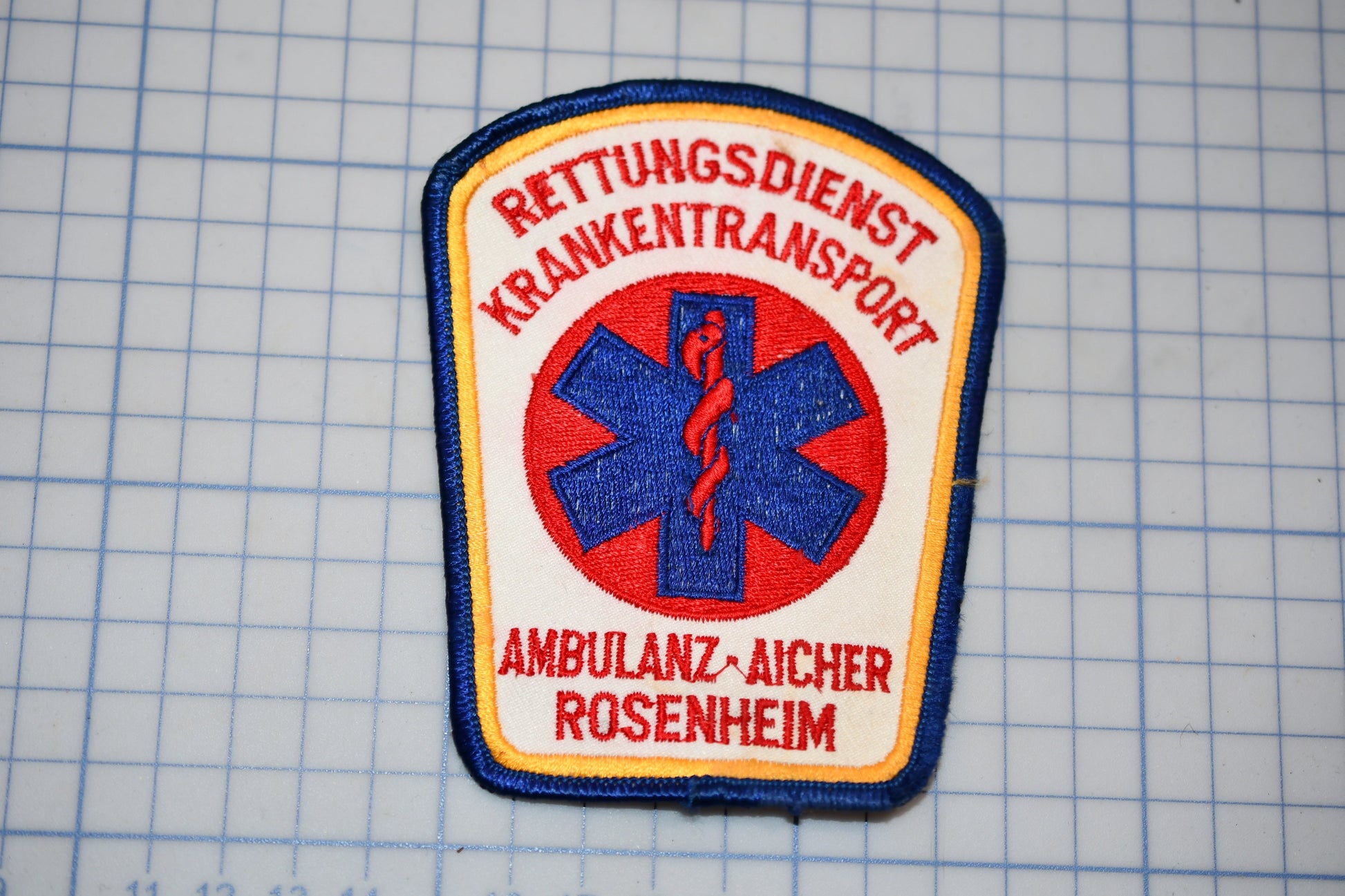 a patch with the text "Rettungsdienst Krankenttransport" and "Ambulanz-Aicher Rosenheim" printed on it, along with a medical symbol.