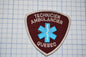 a red and blue patch with the text "TECHNICIEN AMBULANCIER QUEBEC" and a blue medical symbol, indicating that it is a patch for a technician ambulance from Quebec.