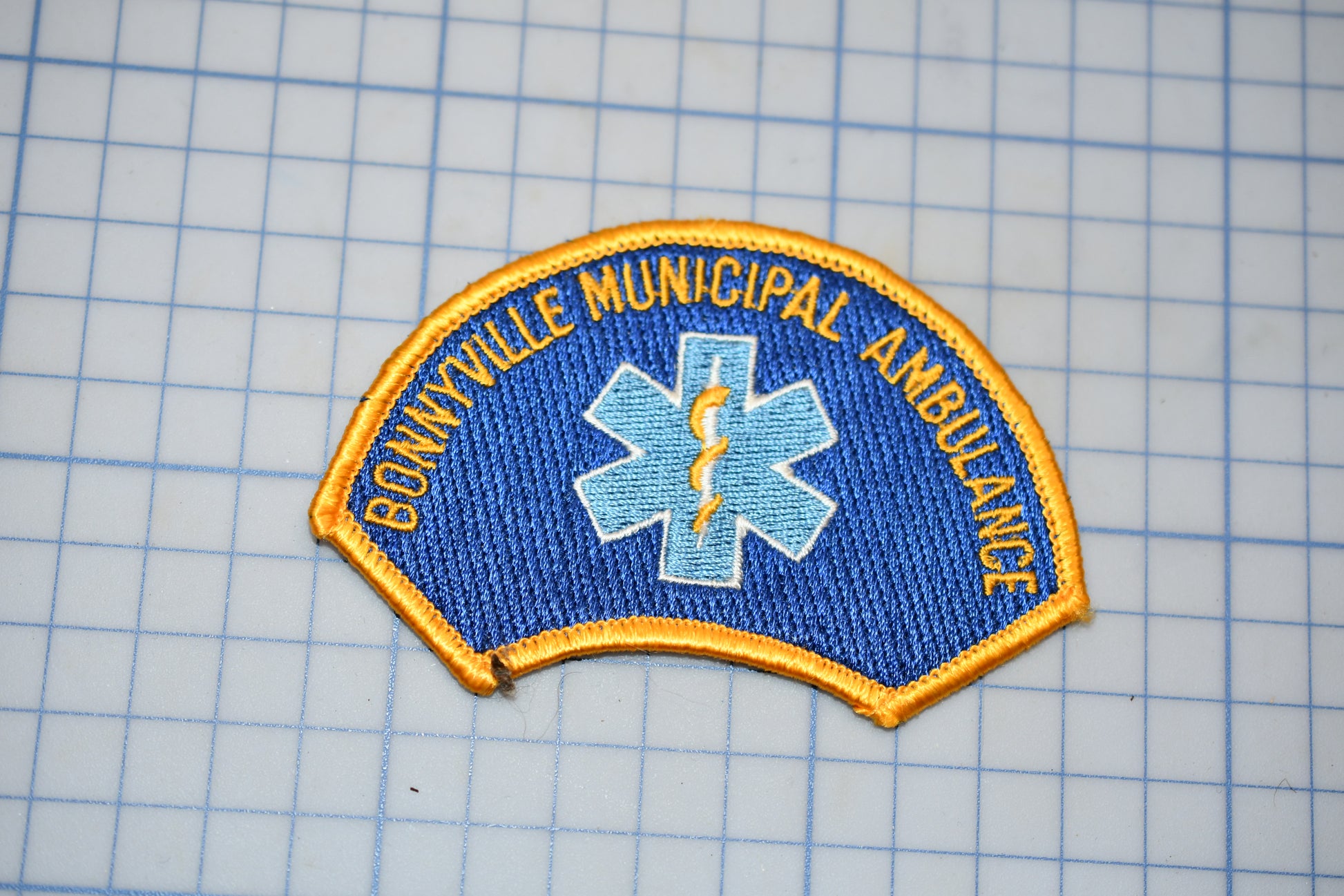 A blue and gold badge with the text "BONNVILLE MUNICIPAL AMBULANCE" and a medical symbol, indicating it is a badge for the Bonnyville municipal ambulance service.